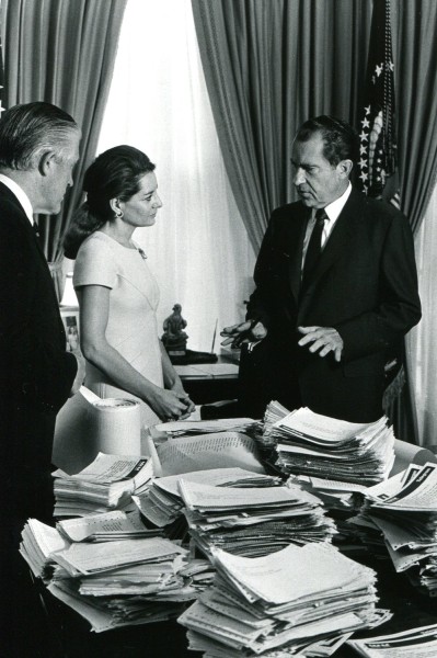 Black-and-white photograph of three people standing around a desk covered in tall stacks of telegrams and documents, with draped curtains and a flag visible behind them