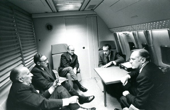 Black-and-white photograph of five men in suits seated in a curved aircraft cabin, mid-conversation, with a Presidential seal visible on the wall