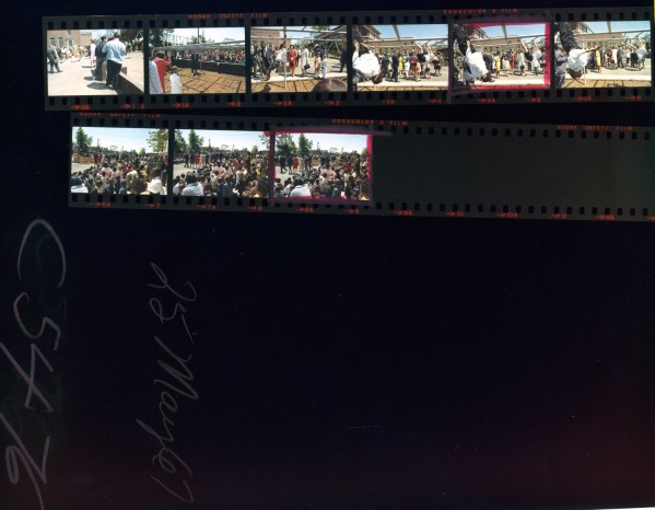 Color contact sheet with two strips of film frames showing large outdoor crowds gathered around playground equipment beside a brick school building on a sunny day