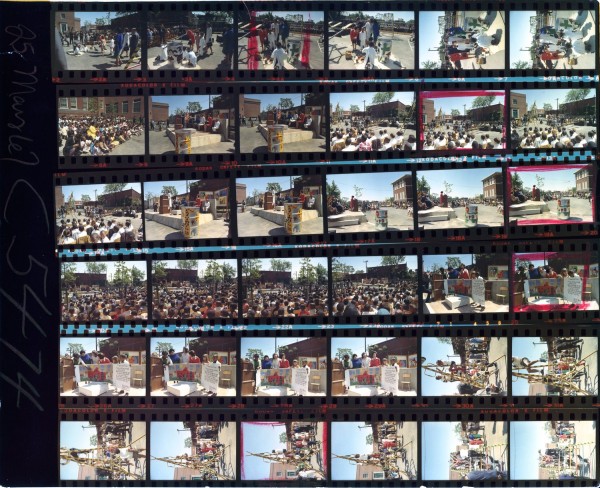 Color contact sheet of approximately 35 film frames showing an outdoor public event with a large crowd, a stage or platform, painted murals, and playground equipment under construction