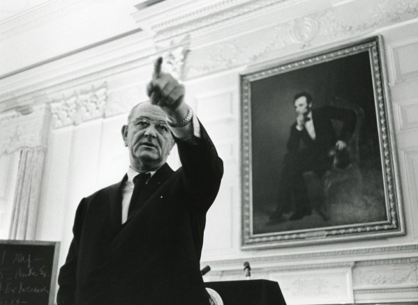 Black-and-white photograph of a man in a dark suit pointing upward, with a large framed portrait of a seated figure hanging on an ornate white wall behind him