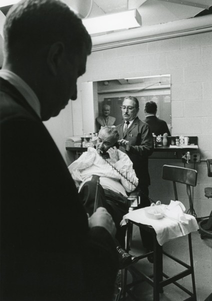 Black-and-white photograph of a man in a barber's cape seated in a chair, holding a telephone handset to his ear while a barber tends to his hair