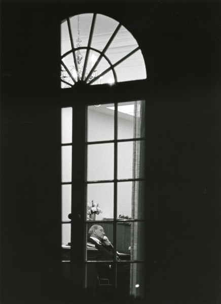 Black-and-white photograph of a man in profile at a desk speaking on a telephone, seen through the tall arched window of a dimly lit exterior