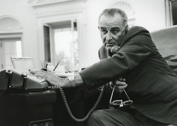 Black-and-white photograph of a man in a dark suit leaning forward, holding a telephone handset to his ear and resting his other hand on a multi-line phone console, eyeglasses in hand