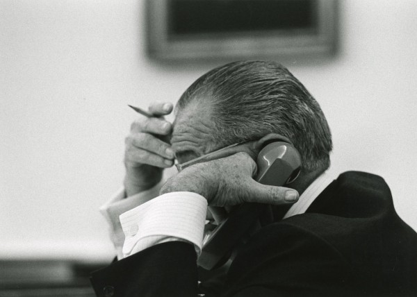 Black-and-white photograph of a man in profile, holding a telephone receiver to his ear with one hand and a pencil near his head with the other