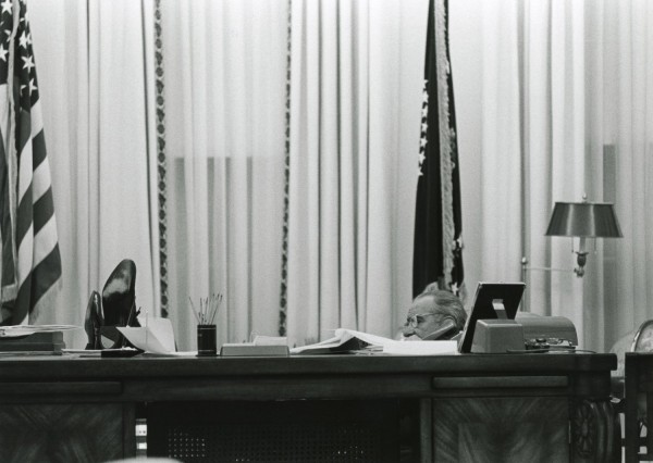 Black-and-white photograph of a man in glasses seated behind a large desk covered with papers and office items, holding a phone to his ear, flanked by flags