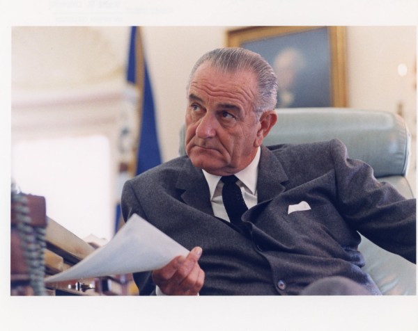 Color photograph of a man in a dark suit seated in a leather chair, holding a document, with a blue flag and a framed portrait visible behind him
