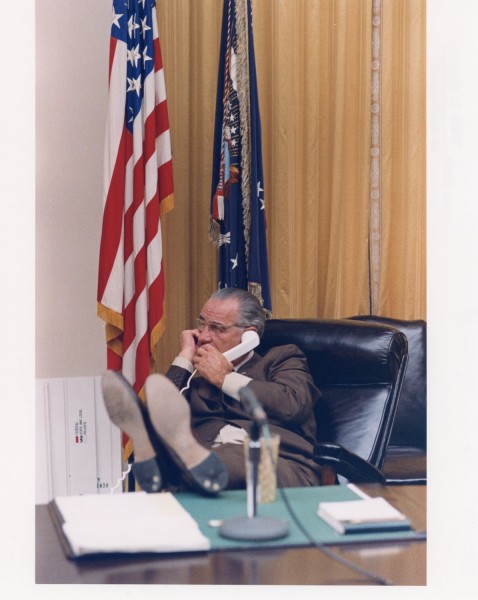 Color photograph of a man in a suit seated in a leather chair, talking on a telephone with feet propped on a large desk. U.S. and presidential flags stand behind him.