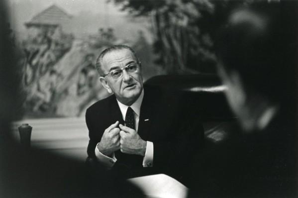 Black-and-white photograph of a man in a dark suit and glasses seated at a table, gesturing with clasped hands while speaking to a person seen from behind in the foreground