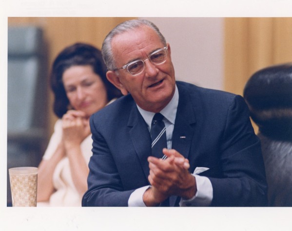 Color photographic print of a man in a dark suit and striped tie, hands clasped, seated at a table; a woman in a light outfit sits softly out of focus behind him