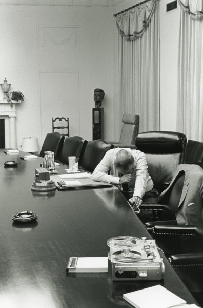 Black-and-white photograph of a man in a white shirt seated alone at a long conference table, head bowed into his hands, reel-to-reel recorder and ashtrays on the table