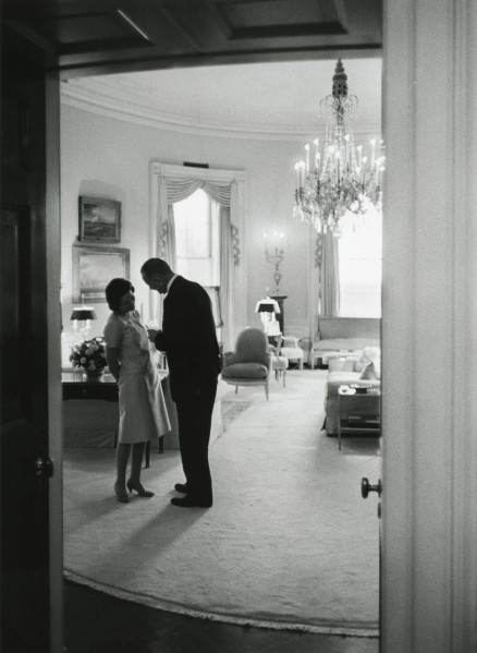 Black-and-white photograph of a man and woman standing close together in a large, elegantly furnished room, shot through a partially open doorway. A chandelier hangs overhead and tall draped windows line the far wall.