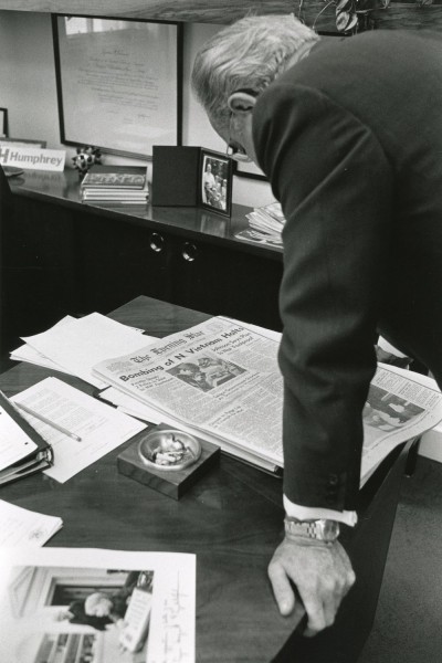 Black-and-white photograph of a man in a dark suit leaning over a paper-strewn desk to read a copy of "The Evening Star" with the headline "Bombing of N. Vietnam Halts"