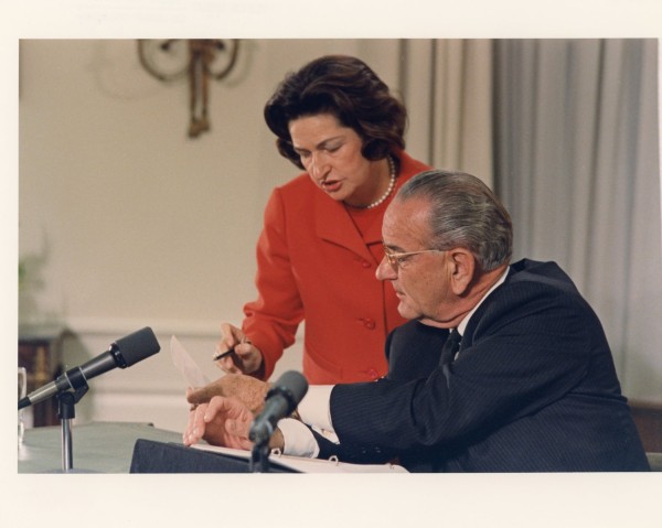 Color photographic print of a woman in a red jacket leaning over a seated man in a dark suit at a table with microphones, both reviewing papers