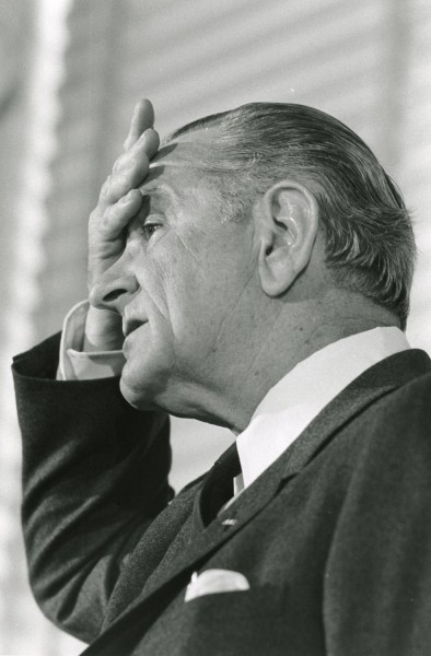Black-and-white photograph of a man in profile, hand raised to his brow, wearing a dark suit and tie