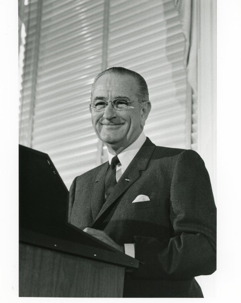 Black-and-white photograph of a smiling older man in a dark suit standing at a wooden podium, with window blinds visible behind him