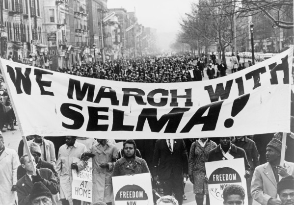 Black-and-white photograph of a large street march. Marchers carry a wide banner reading "We March With Selma!" and signs reading "Freedom Now" as crowds fill the street behind them